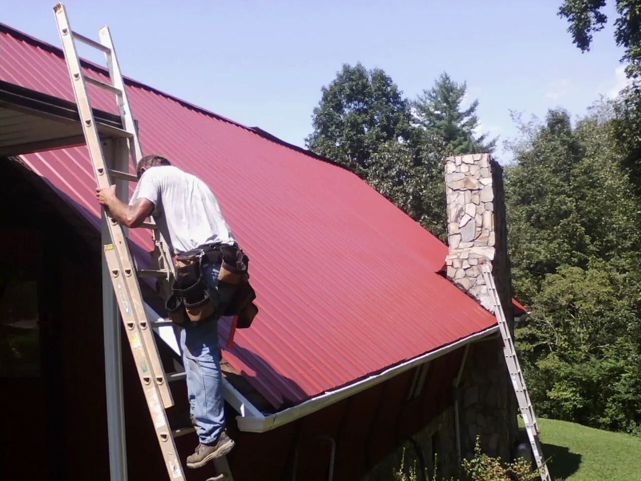 Roofer on a ladder beside a red metal roof and stone chimney — Soffit & Fascia Repair by Roofing Brothers in Pocono Summit